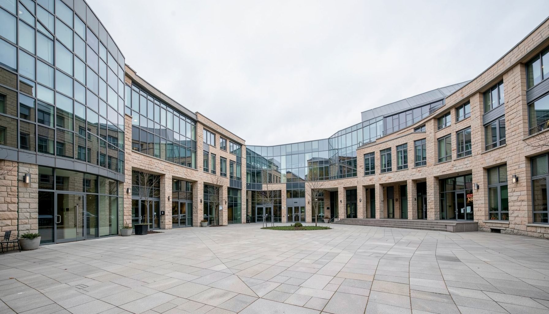 Wide-angle view of a modern university campus courtyard with glass and stone buildings under a soft overcast sky, calm academic atmosphere